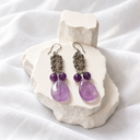 A pair of dangling earrings with ornate silver-toned filigree tops, small purple beads, and large teardrop-shaped purple amethyst drops, displayed on white textured stone blocks over white fabric.
