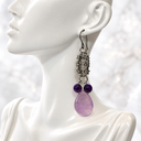 A single dangling earring displayed on a white mannequin bust. The earring has an oxidized silver-toned metal chain and hook, with two small dark purple beads and a larger teardrop-shaped light purple stone at the bottom.