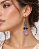 A close-up of a woman wearing a dangling earring with an ornate dark metal filigree top, two small dark purple beads, and a large teardrop-shaped light purple stone.